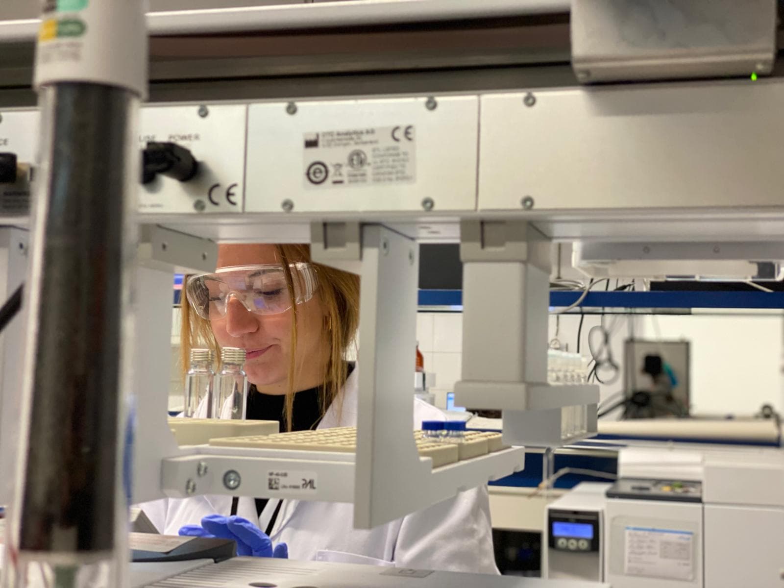 Una investigadora amb bates de laboratori i ulleres de seguretat mira atentament una mostra en una safata de vials en un entorn de laboratori científic