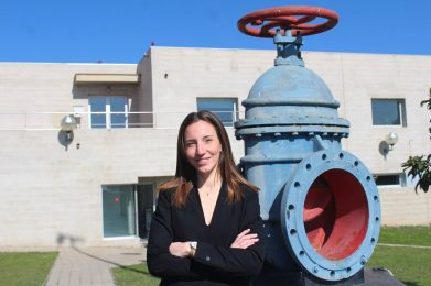 Una dona professional somrient, amb un vestit jaqueta negre i els braços creuats, de peu davant d'una gran vàlvula industrial blava a l'exterior de l'oficina.